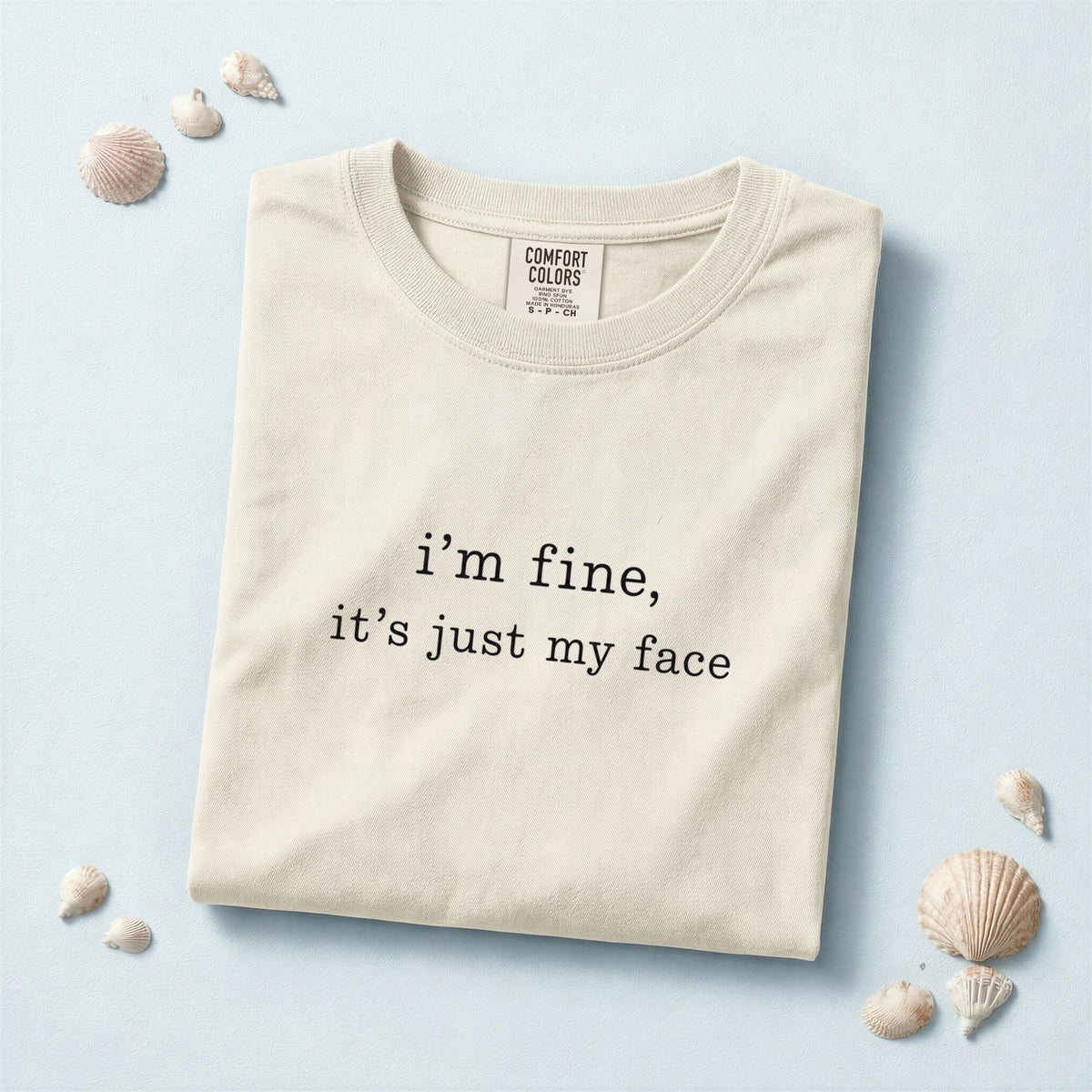May include: A cream-colored short-sleeve t-shirt featuring the black printed text "i'm fine, it's just my face" centered on the chest. The shirt has a crew neckline and a Comfort Colors brand label visible on the inside of the collar area.