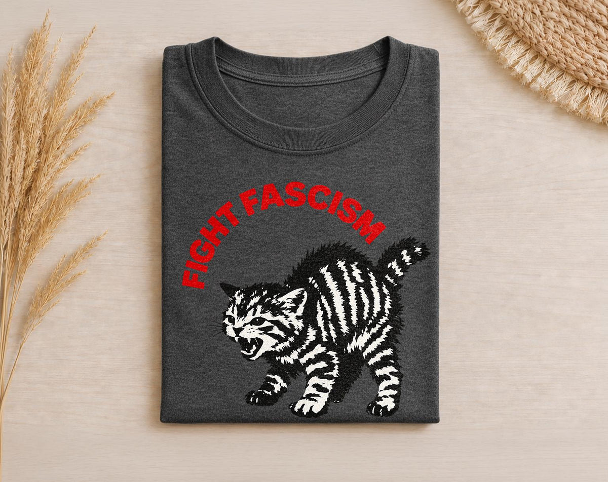 May include: A dark charcoal gray t-shirt featuring a graphic of a hissing, arched-back tabby kitten in black and white. Above the kitten, the text FIGHT FASCISM is printed in a bold, red, sans-serif font, following a slight upward curve.