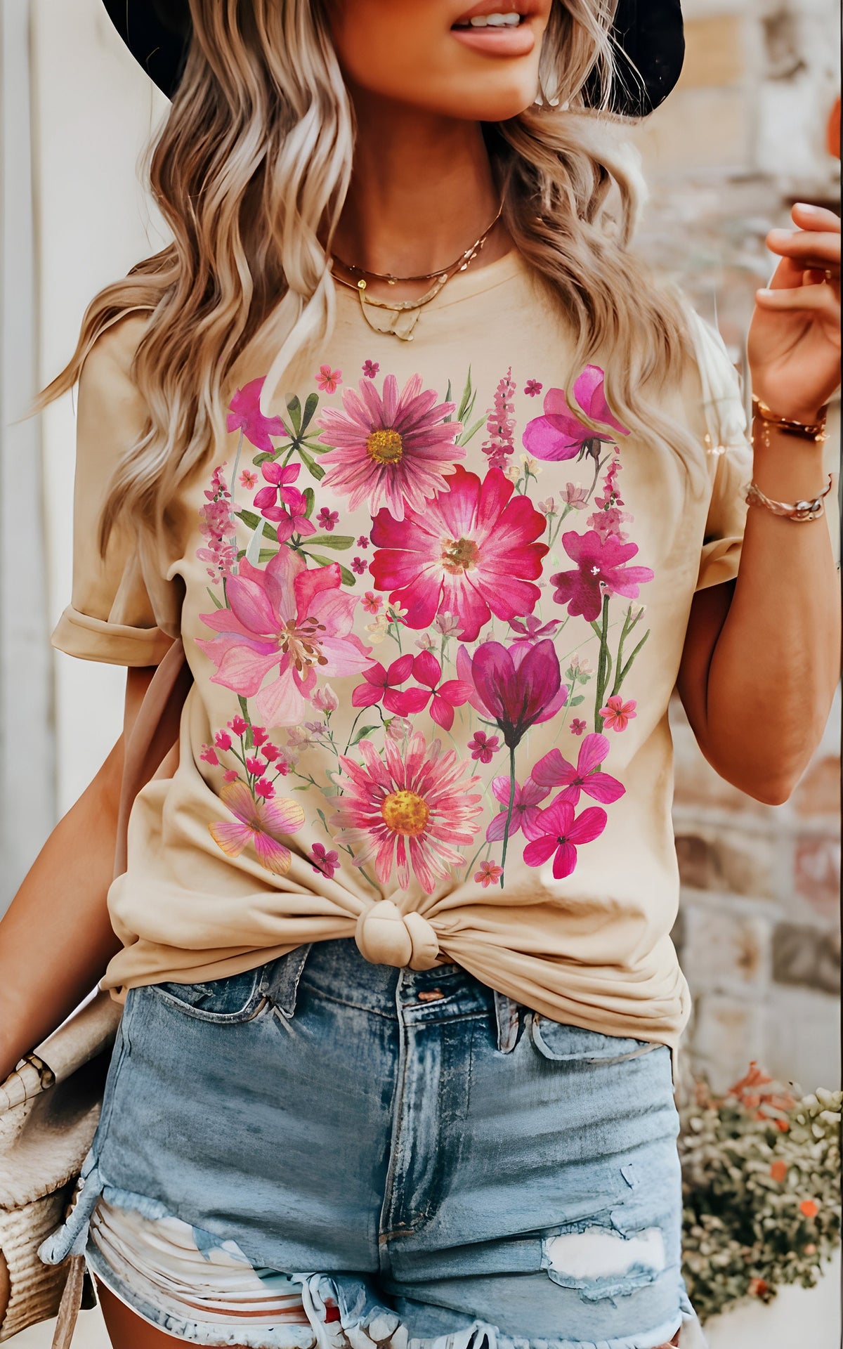May include: Beige t-shirt featuring a vibrant floral design in shades of pink and magenta. The flowers are arranged in a clustered pattern across the front. The shirt is knotted at the waist and paired with denim shorts.