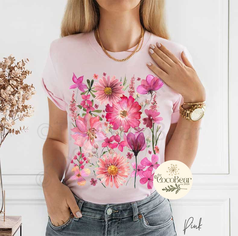 May include: A light pink t-shirt featuring a watercolor floral design in shades of pink and magenta. The design includes various flowers and foliage. The text "Pink" is at the bottom right.