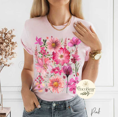 May include: A light pink t-shirt featuring a watercolor floral design in shades of pink and magenta. The design includes various flowers and foliage. The text "Pink" is at the bottom right.