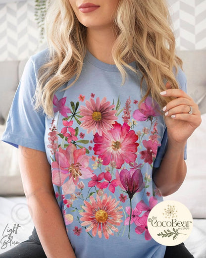 May include: Light blue t-shirt featuring a vibrant floral design in shades of pink and red. The design includes various flower types, creating a botanical theme. The text "Light Blue" is visible on the left side of the shirt. The CocoBear logo is on the right.