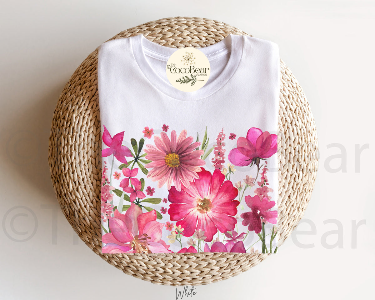 May include: White t-shirt featuring a vibrant floral design with pink and red flowers, green leaves, and the text "White" at the bottom. The shirt is displayed on a woven, circular surface, with a tag that reads "CocoBear Design".