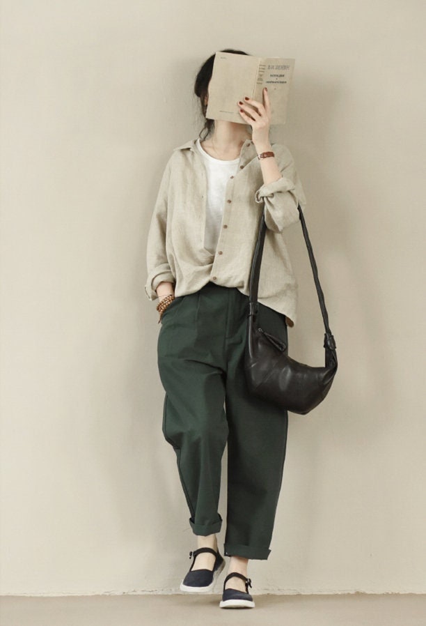 May include: A woman wearing a beige linen shirt, dark green pants, and black leather shoes. She is carrying a black leather shoulder bag and holding a book in front of her face.