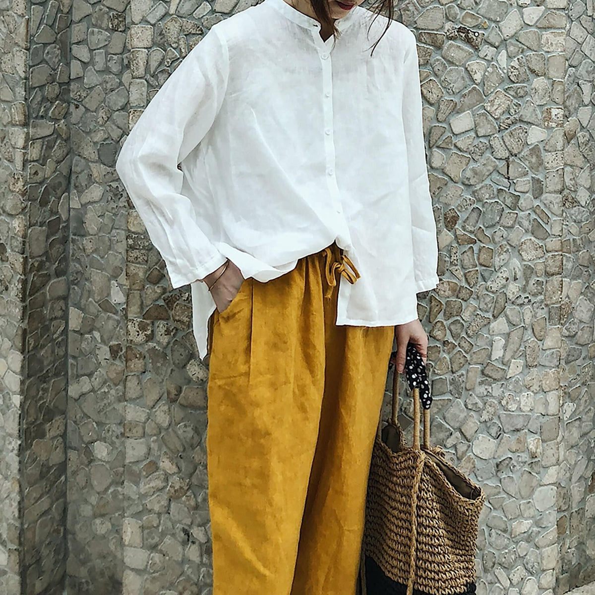 May include: A white linen shirt with a mandarin collar and long sleeves, paired with mustard yellow linen pants and a woven straw tote bag with black polka dot handles.