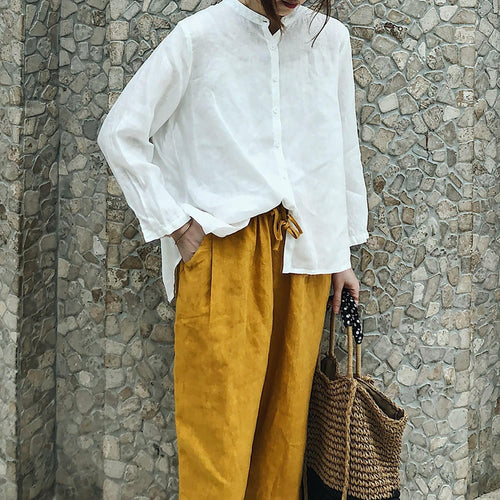May include: A white linen shirt with a mandarin collar and long sleeves, paired with mustard yellow linen pants and a woven straw tote bag with black polka dot handles.
