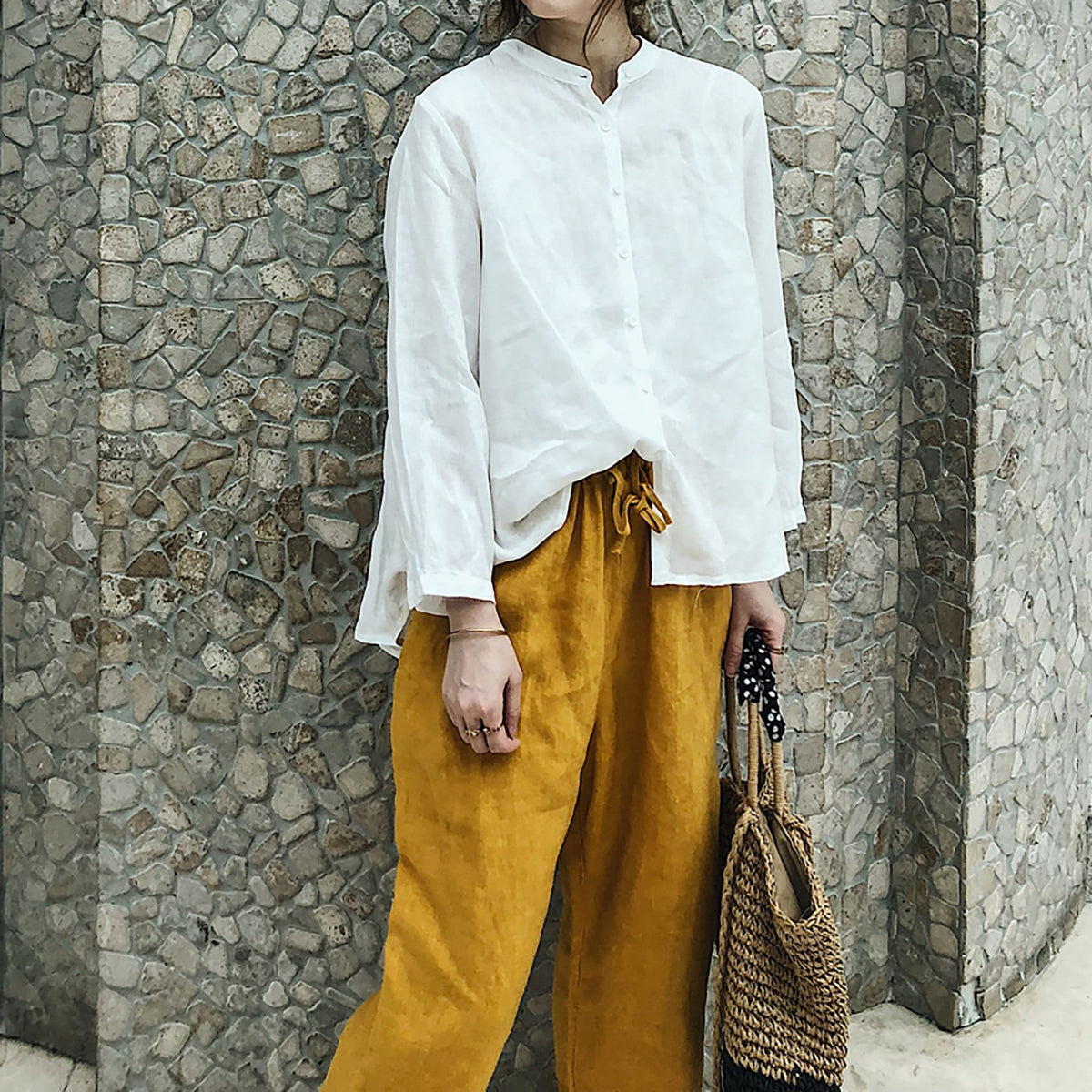 May include: A white linen shirt with a mandarin collar and long sleeves, paired with mustard yellow linen pants and a woven straw tote bag with black accents.