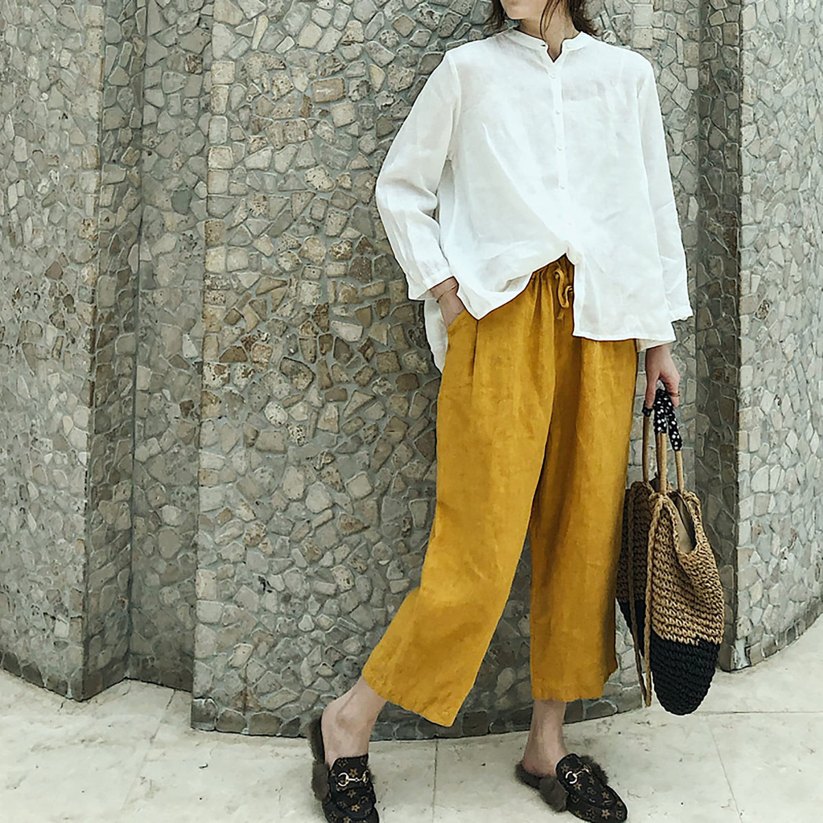 May include: A woman wearing a white linen shirt with a mandarin collar and long sleeves, paired with mustard yellow wide-leg cropped pants. She is carrying a woven straw tote bag with black accents.