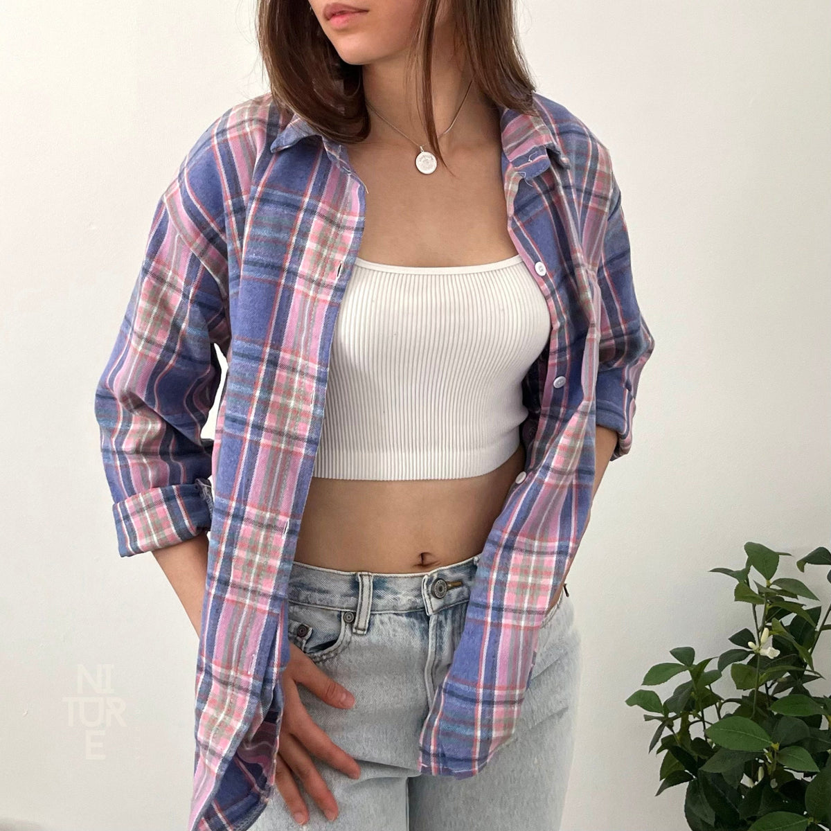 May include: A blue and pink plaid flannel shirt with a white tank top underneath. The shirt is unbuttoned and the tank top is visible. The person is wearing blue jeans.
