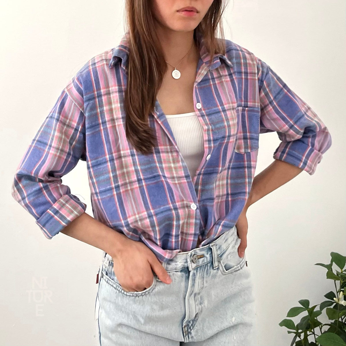 May include: A woman wearing a blue and pink plaid flannel shirt with a white tank top underneath. She is wearing light blue denim jeans and has her hand in her pocket.