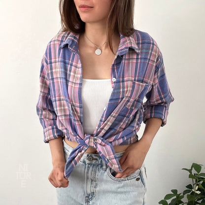 May include: A blue and pink plaid flannel shirt tied at the waist over a white tank top and light blue denim jeans.