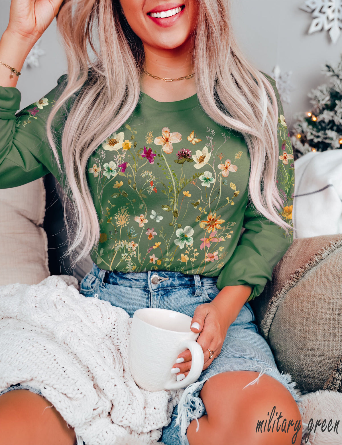 May include: A woman wearing a long-sleeved, military green shirt with a floral print. She is holding a white mug and sitting on a couch with a white knitted blanket.