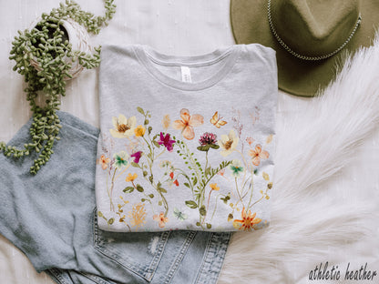 May include: A gray t-shirt with a watercolor floral print featuring yellow, pink, and purple flowers. The shirt is folded and laid on a white fuzzy surface with a pair of blue jeans and a green hat.