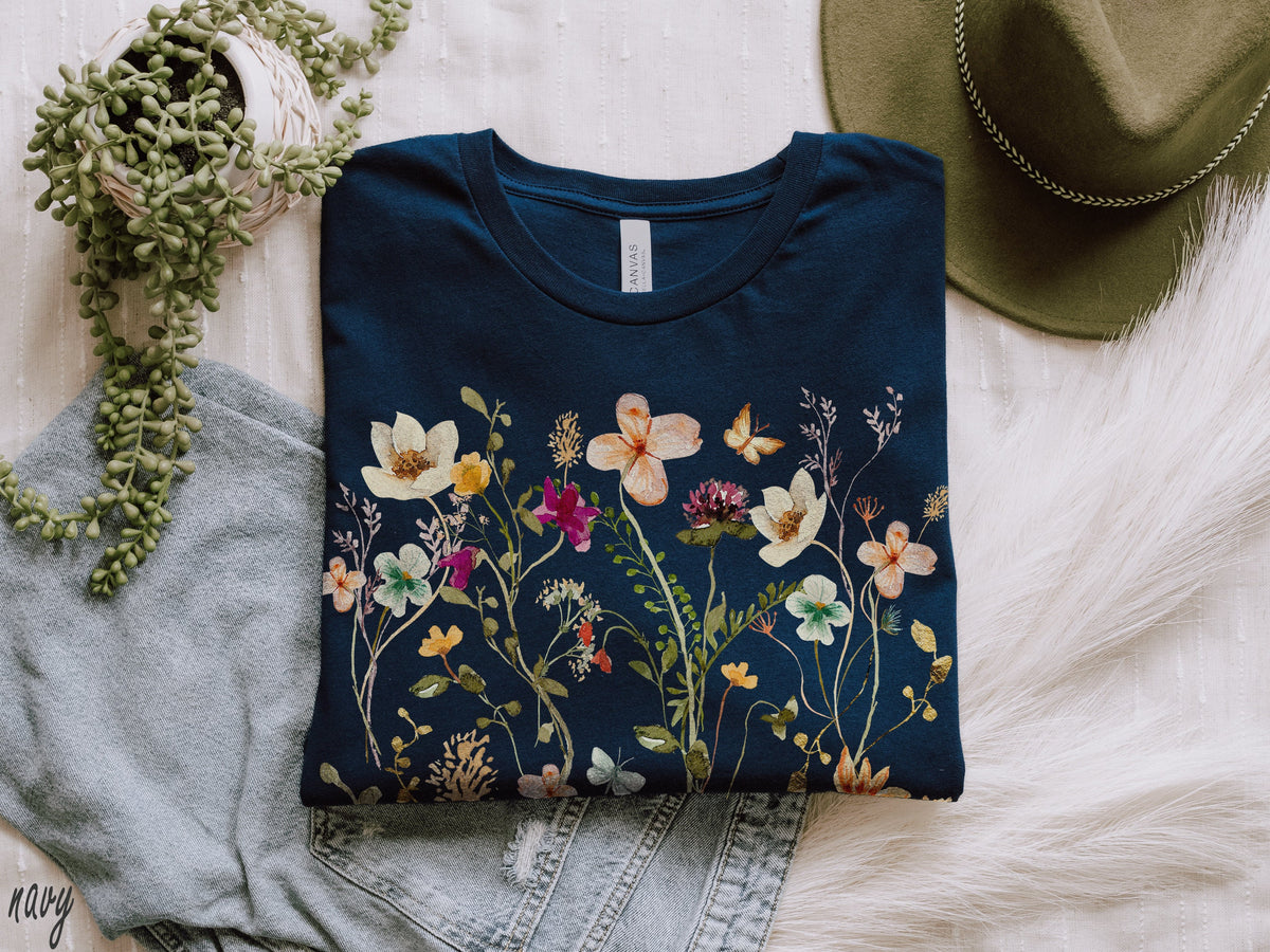 May include: A navy blue t-shirt with a floral print featuring a variety of flowers and butterflies. The shirt is folded and laid on a white surface.