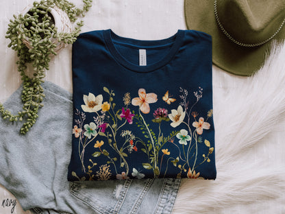 May include: A navy blue t-shirt with a floral print featuring a variety of flowers and butterflies. The shirt is folded and laid on a white surface.