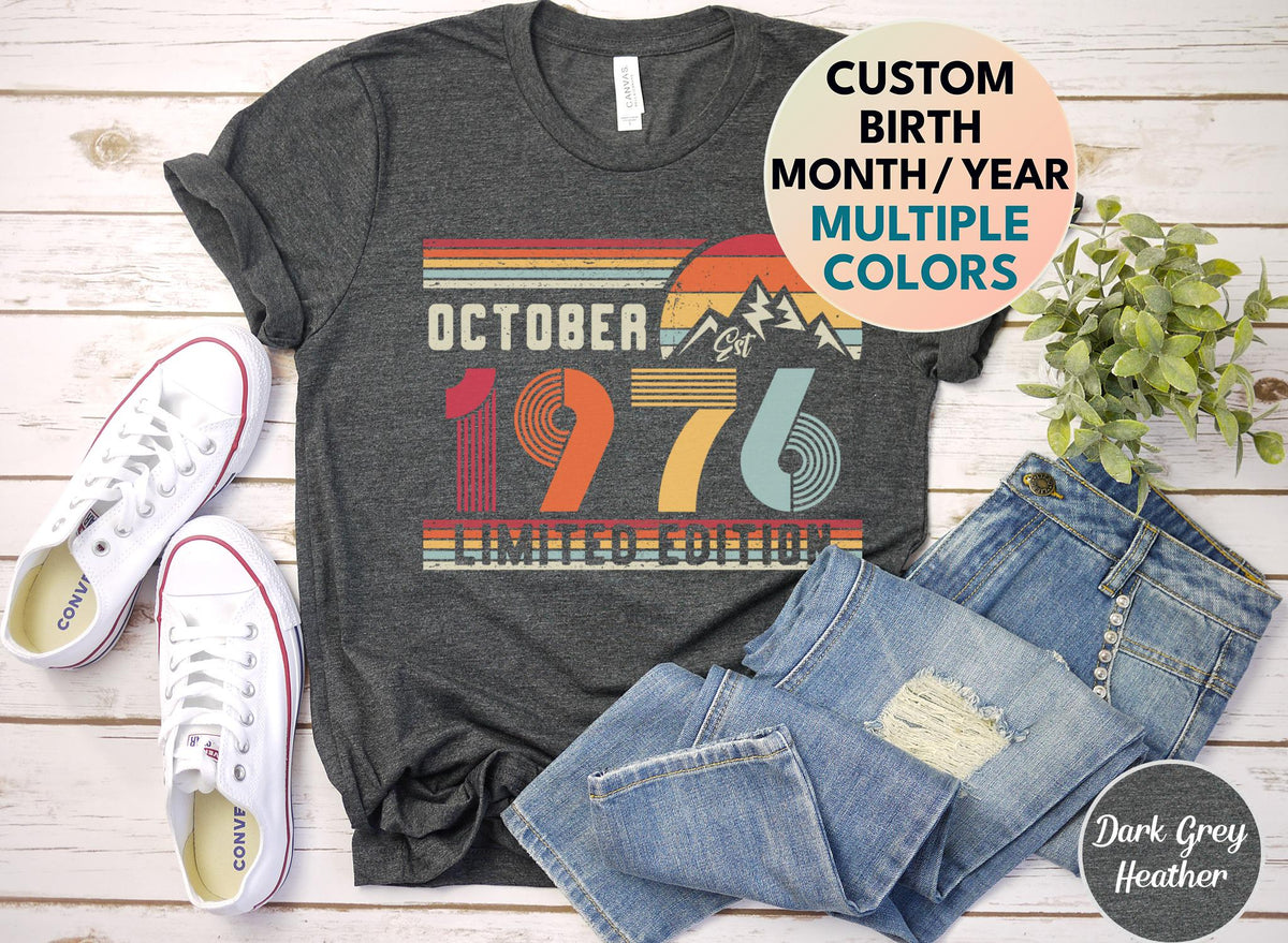 May include: A dark grey heather t-shirt featuring a vintage-style graphic with the text OCTOBER 1976 LIMITED EDITION, a mountain silhouette, and a sunset design in shades of red, orange, yellow, and blue, with a circular label stating CUSTOM BIRTH MONTH/YEAR MULTIPLE COLORS.