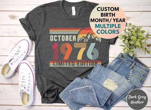 May include: A dark grey heather t-shirt featuring a vintage-style graphic with the text OCTOBER 1976 LIMITED EDITION, a mountain silhouette, and a sunset design in shades of red, orange, yellow, and blue, with a circular label stating CUSTOM BIRTH MONTH/YEAR MULTIPLE COLORS.