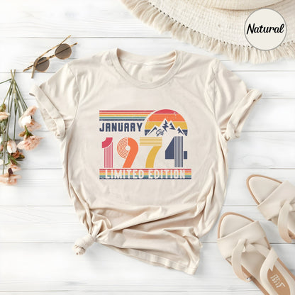 May include: A natural-colored t-shirt featuring a retro-style graphic with the text "JANUARY 1974 LIMITED EDITION". The design includes a sunset, mountain peaks, and horizontal stripes in red, orange, yellow, and blue, printed on the front of the shirt.