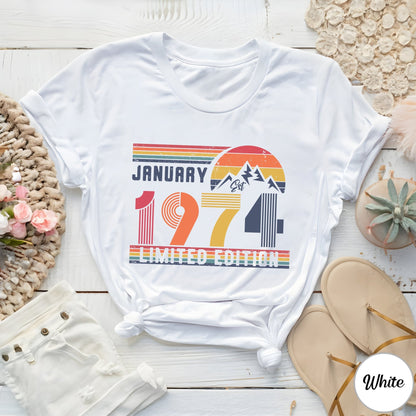 May include: A white short-sleeved t-shirt featuring a graphic print with the text "JANUARY 1974 LIMITED EDITION". The design includes a retro-style sunset, mountain silhouettes, and horizontal stripes in shades of red, orange, yellow, and blue.