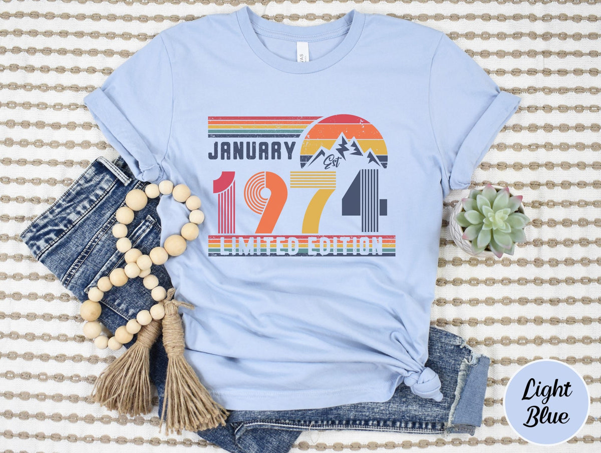 May include: A light blue t-shirt featuring a graphic print with the text JANUARY 1974 LIMITED EDITION. The design includes a retro-style sunset, mountain silhouette, and horizontal stripes in red, orange, yellow, and blue colors across the chest.