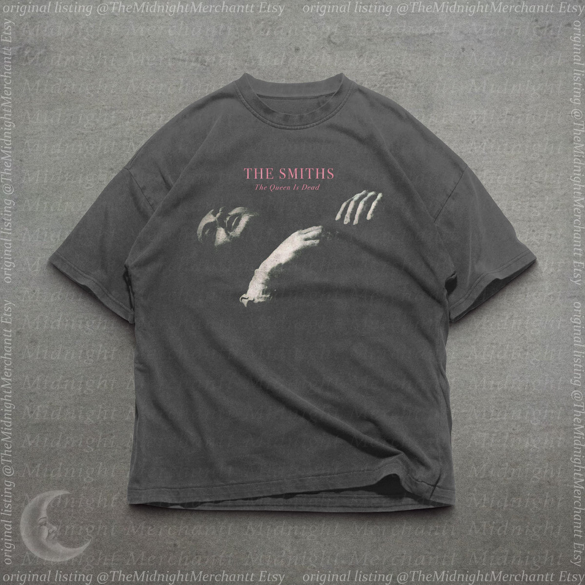 May include: A charcoal gray short-sleeve t-shirt featuring a graphic print of a person's face and hand. The text "THE SMITHS" appears in pink above the words "The Queen Is Dead" in a smaller white font, centered on the chest of the garment.