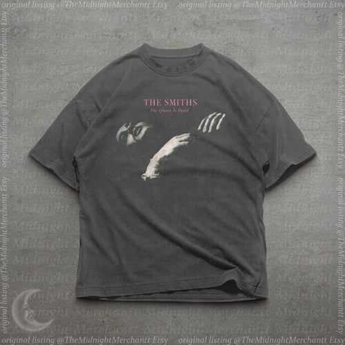 May include: A charcoal gray short-sleeve t-shirt featuring a graphic print of a person's face and hand. The text "THE SMITHS" appears in pink above the words "The Queen Is Dead" in a smaller white font, centered on the chest of the garment.