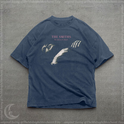 May include: A dark blue short-sleeve t-shirt featuring a graphic print of a person's face and hand. The text THE SMITHS and The Queen Is Dead is printed in pink and white above the graphic. The shirt has a crew neckline and a relaxed fit.