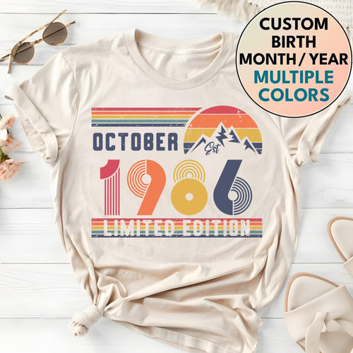 May include: A cream-colored t-shirt featuring a graphic print with the text OCTOBER 1986 LIMITED EDITION. The design includes a retro-style sunset, mountain silhouette, and horizontal stripes in shades of red, orange, yellow, and blue.