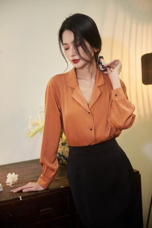 May include: A woman wearing a long-sleeved, rust-colored blouse with a button-down front and a black skirt. She is standing in front of a wooden dresser with a white flower on it.