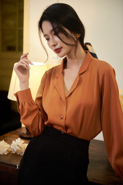 May include: A woman wearing a long-sleeved, rust-colored blouse with a button-up front and a black skirt. She is standing in front of a wooden table with a lamp and flowers.