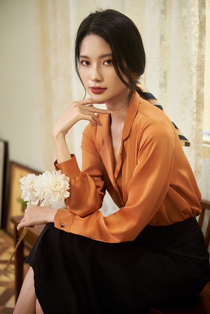 May include: A woman wearing a burnt orange long-sleeved blouse with a black skirt and a striped scarf tied around her hair. She is holding a bouquet of white flowers.