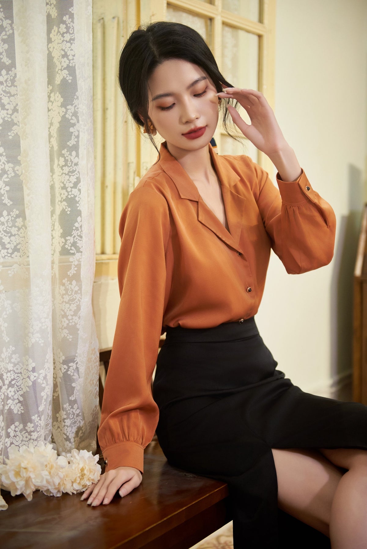 May include: A woman wearing a rust-colored long-sleeved blouse with a black skirt. The skirt has a slit on the side. She is sitting on a wooden bench.