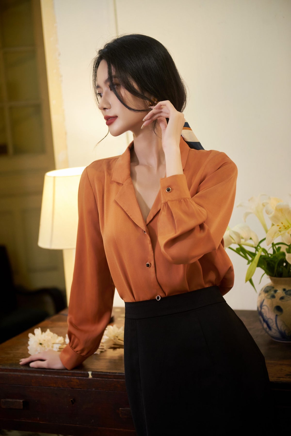 May include: A woman wearing a rust-colored long-sleeved blouse with a button-up front and a black skirt. She is standing in front of a wooden table with a lamp and a vase of white flowers.