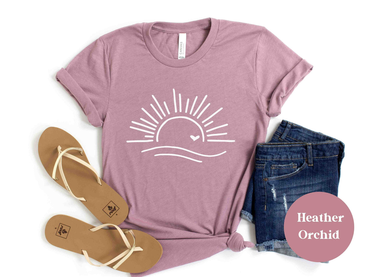 May include: A heather orchid t-shirt with a white graphic of a sun with a heart over a wave. The shirt is laid flat on a white background with a pair of brown sandals and denim shorts.