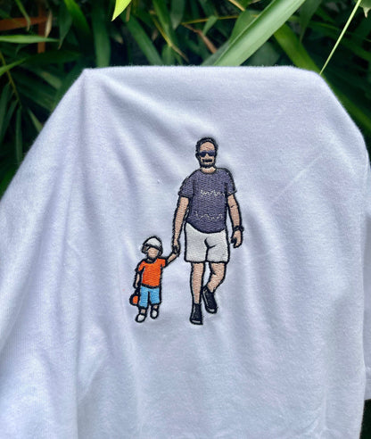 May include: A white t-shirt featuring an embroidered graphic of an adult and a small child walking while holding hands. The adult wears a dark purple t-shirt, white shorts, and sunglasses. The child wears an orange shirt, blue shorts, and a white hat.