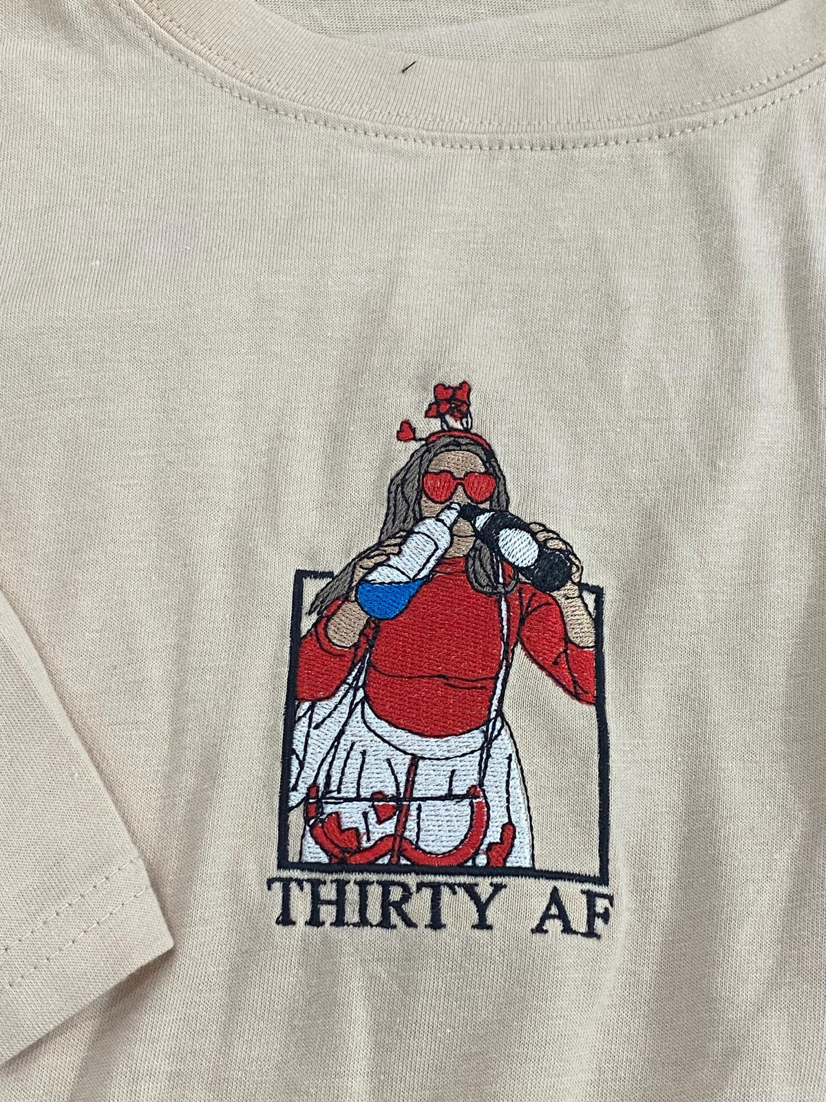 May include: A beige t-shirt featuring an embroidered graphic of a figure wearing heart-shaped sunglasses and a headband, drinking from two bottles. The figure wears a red top and white skirt, framed in a black box with the text THIRTY AF below.