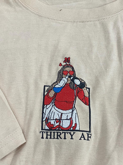 May include: A beige t-shirt featuring an embroidered graphic of a figure wearing heart-shaped sunglasses and a headband, drinking from two bottles. The figure wears a red top and white skirt, framed in a black box with the text THIRTY AF below.