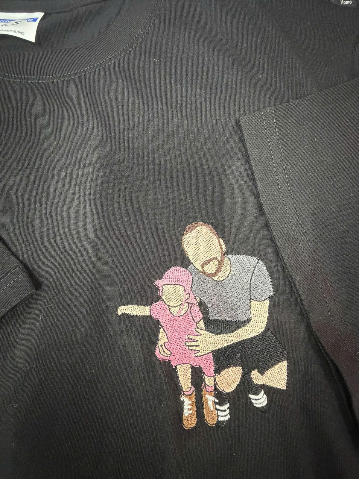 May include: A black t-shirt featuring an embroidered design of a person kneeling while holding a small child. The child wears a pink dress and hat, while the adult wears a gray t-shirt and black shorts. The figures have no facial features.
