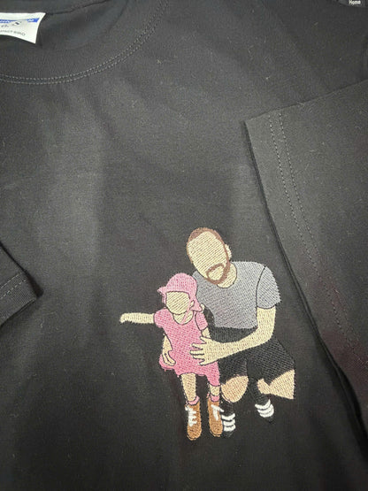 May include: A black t-shirt featuring an embroidered design of a person kneeling while holding a small child. The child wears a pink dress and hat, while the adult wears a gray t-shirt and black shorts. The figures have no facial features.