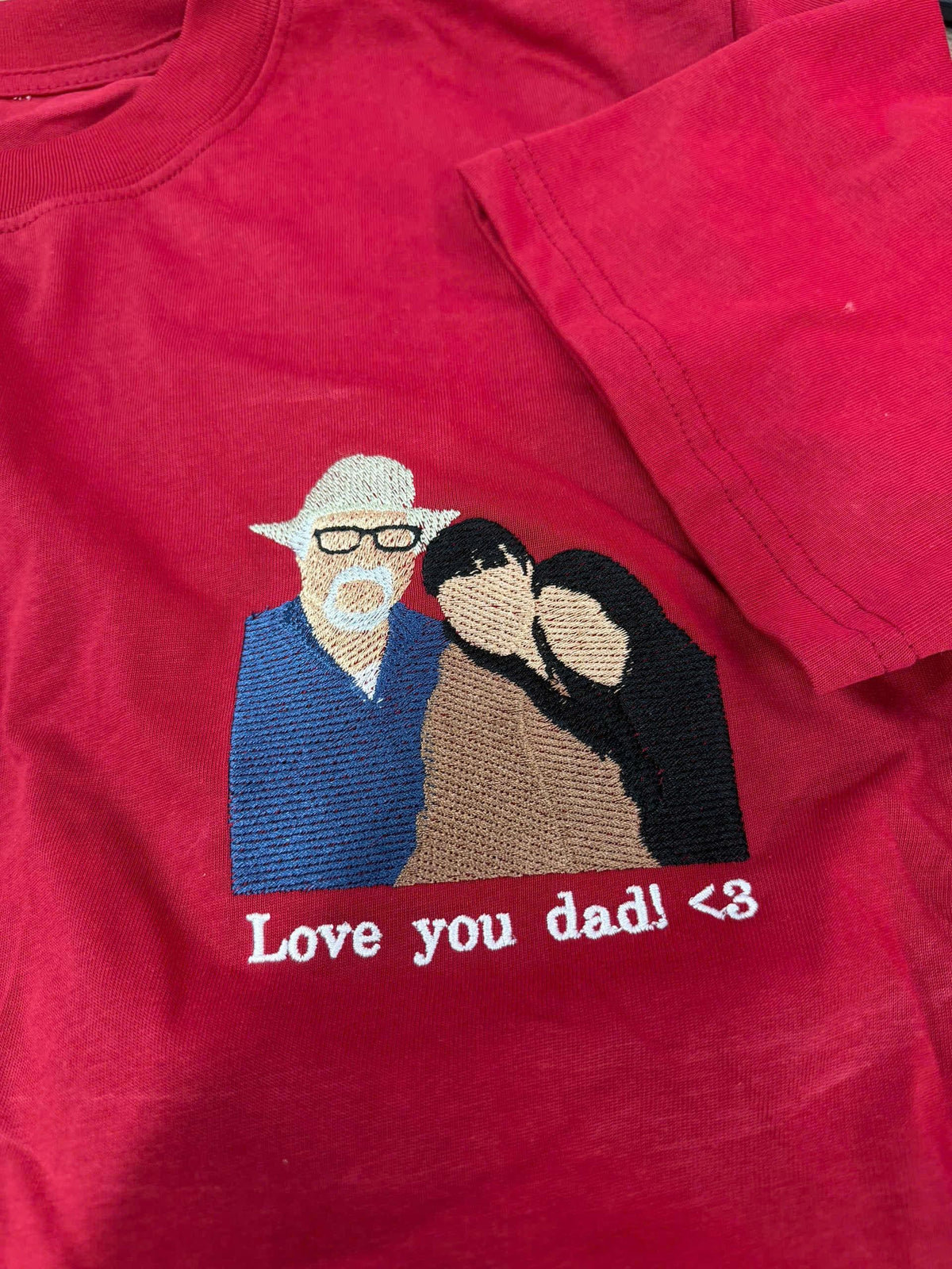 May include: A red t-shirt featuring an embroidered design of three figures. One figure wears a white hat and glasses, while the others are positioned beside them. Below the embroidery, the white text reads: Love you dad! <3