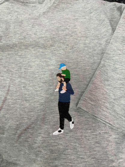 May include: A heather gray t-shirt featuring an embroidered design of a person with a beard wearing a dark blue long-sleeve shirt and black pants, carrying a small child on their shoulders who wears a green shirt and a bright blue baseball cap.
