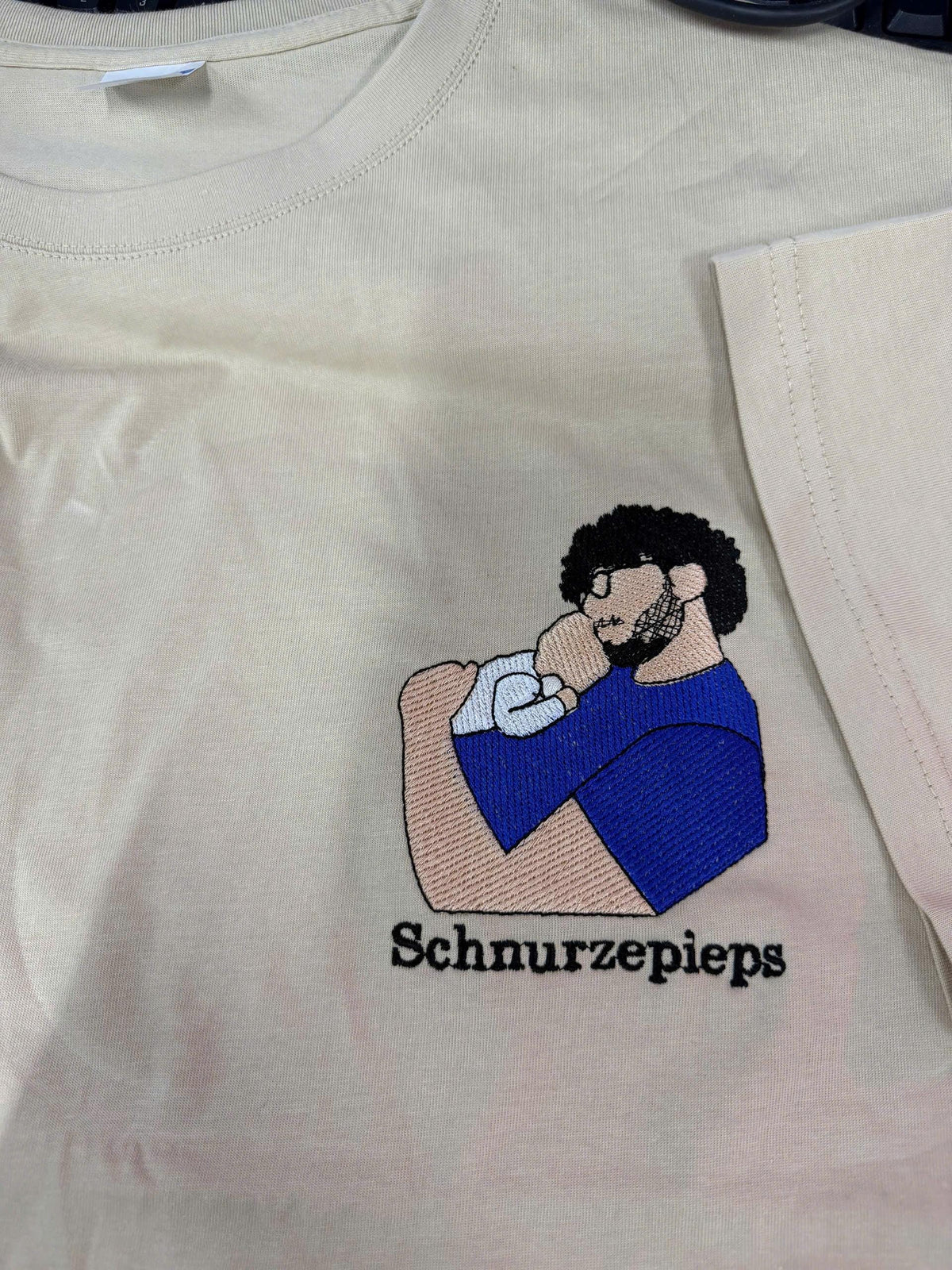 May include: A cream-colored t-shirt featuring an embroidered graphic of a person with dark curly hair, glasses, and a beard holding a baby. The person wears a blue shirt. The text "Schnurzepieps" is embroidered in black below the graphic.