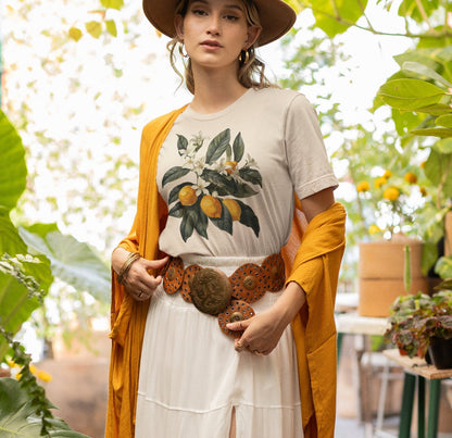 May include: A woman wearing a white t-shirt with a lemon tree graphic print, a brown hat, a white skirt, and a brown leather belt with a large metal buckle. She is standing in front of a green leafy background.