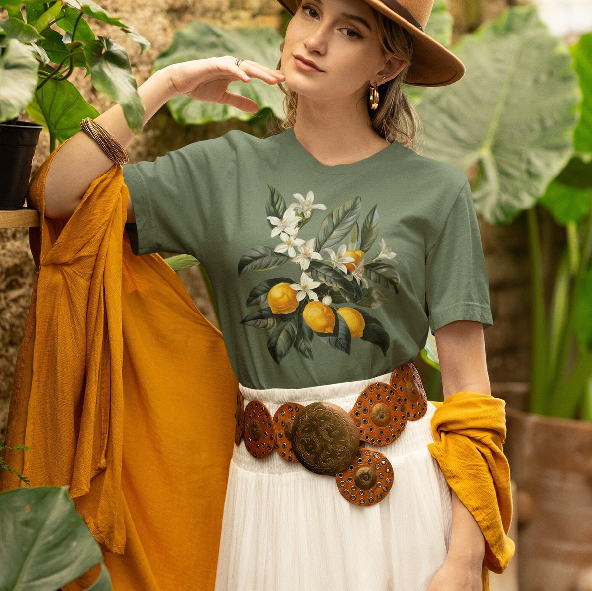 May include: A woman wearing a green t-shirt with a floral lemon print and a brown leather belt with large metal accents. She is wearing a white skirt and a yellow scarf.
