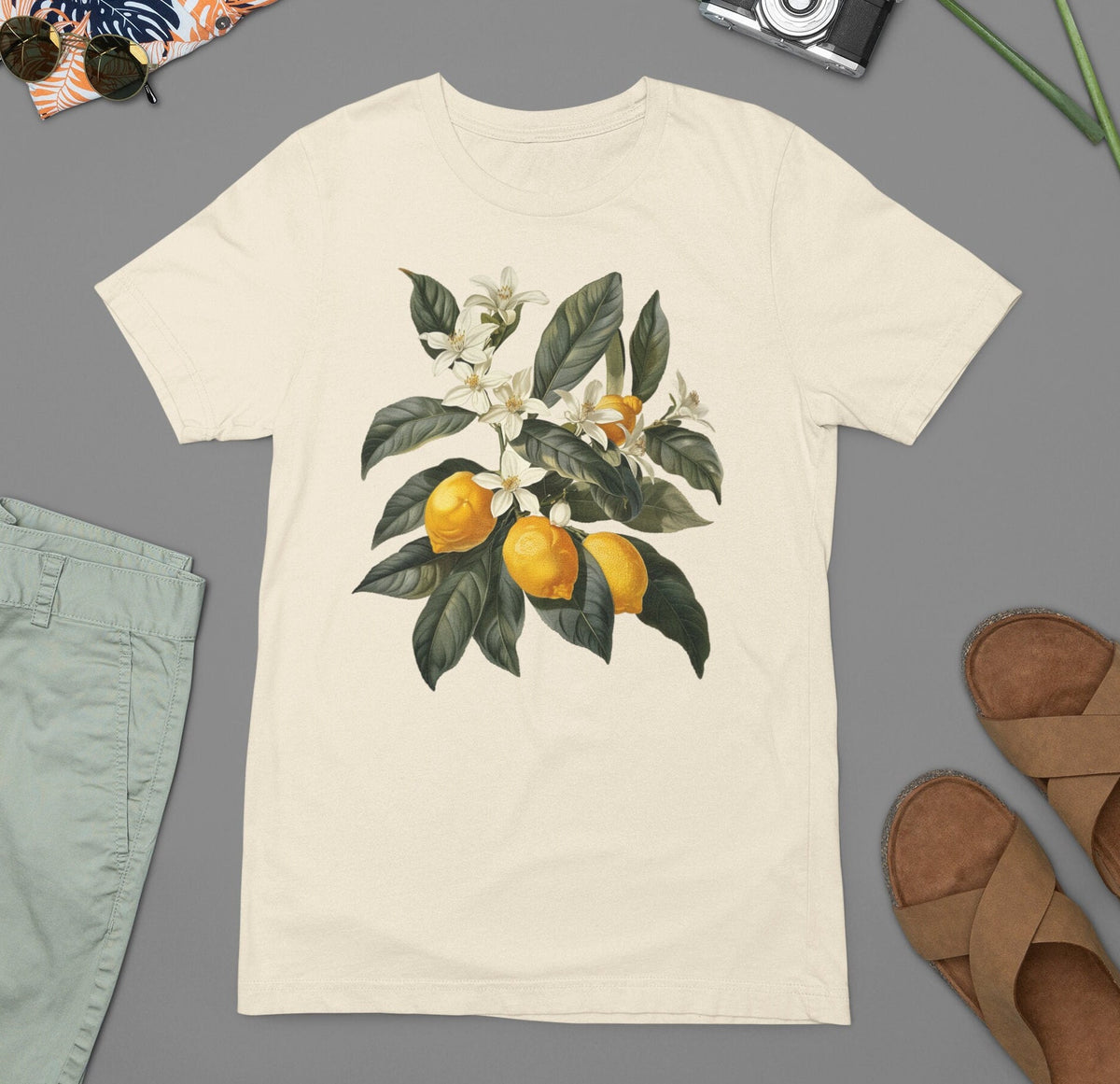 May include: A cream colored t-shirt with a graphic of a lemon tree branch with white blossoms and yellow lemons.