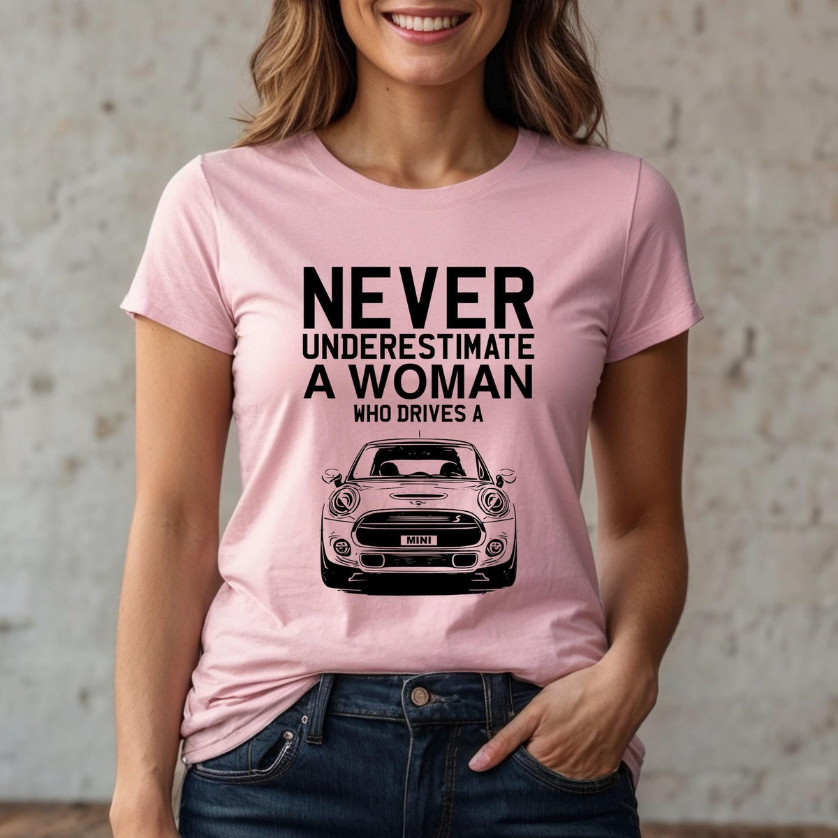 May include: A light pink short-sleeved t-shirt featuring black text that reads NEVER UNDERESTIMATE A WOMAN WHO DRIVES A, positioned above a black line-art illustration of the front view of a Mini Cooper car with the word MINI on the license plate.