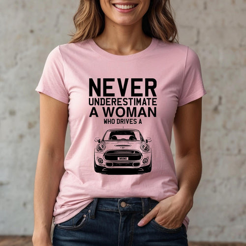 May include: A light pink short-sleeved t-shirt featuring black text that reads NEVER UNDERESTIMATE A WOMAN WHO DRIVES A, positioned above a black line-art illustration of the front view of a Mini Cooper car with the word MINI on the license plate.