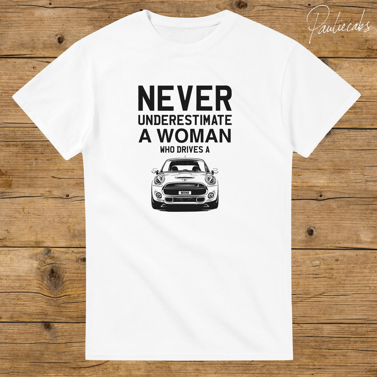 May include: A white short-sleeved t-shirt featuring black text that reads NEVER UNDERESTIMATE A WOMAN WHO DRIVES A above a black and white line art illustration of the front view of a Mini Cooper car with the word MINI on the license plate.