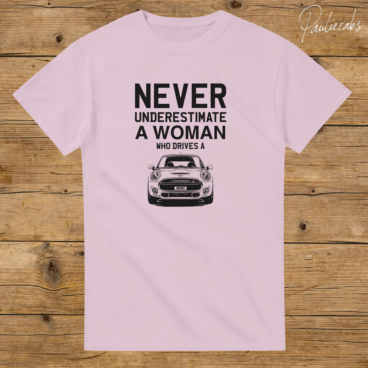 May include: A light pink t-shirt featuring black text that reads NEVER UNDERESTIMATE A WOMAN WHO DRIVES A above a black and white graphic illustration of the front view of a Mini Cooper car with the word MINI on the license plate area.