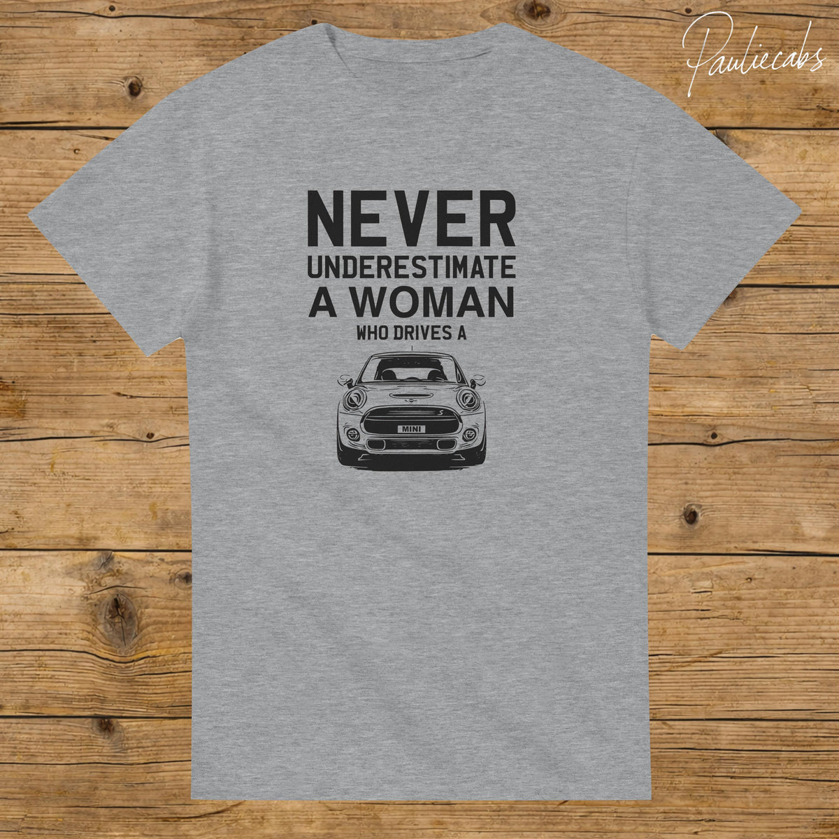 May include: A heather gray short-sleeved t-shirt featuring black text that reads NEVER UNDERESTIMATE A WOMAN WHO DRIVES A, positioned above a black and white graphic illustration of a Mini Cooper car with the word MINI on the license plate.
